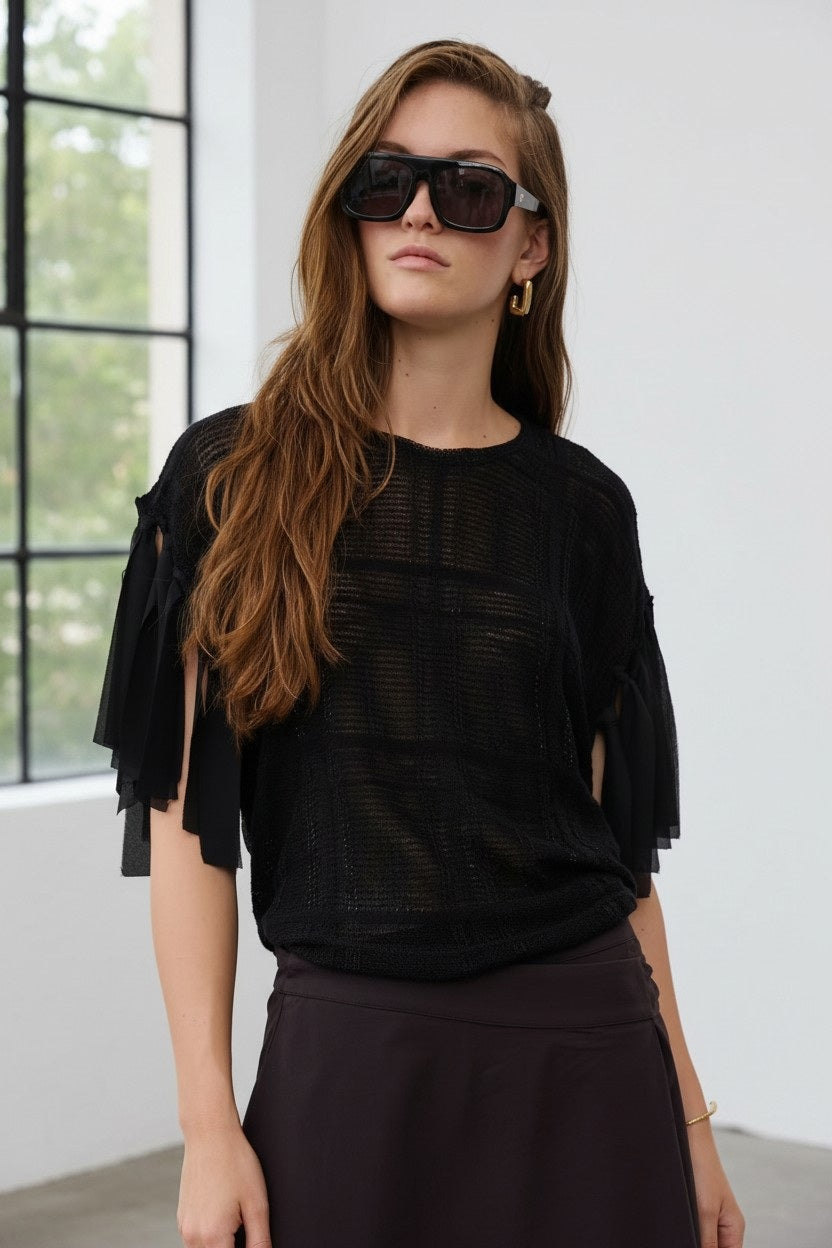 A woman with long brown hair, wearing large black sunglasses and the FLOOR - KNIT FRINGE TOP IN BLACK by FLOOR, featuring an open-knit construction, stands indoors near a window and looks confidently at the camera.
