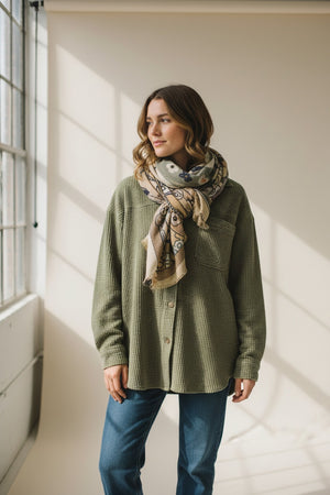 The STORIATIPIC - GAYA Wool Scarf in Beige by STORIATIPIC is a lightweight wool and silk scarf featuring a floral and geometric pattern in muted green, beige, orange, and navy tones, shown loosely knotted against a plain white background.