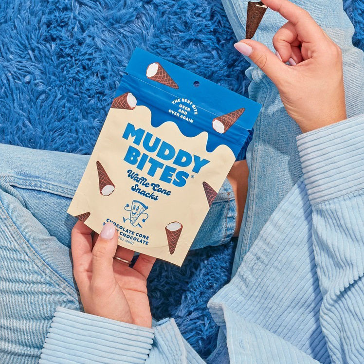 A person dressed in a light blue outfit sits on a plush, royal blue rug holding a bag of Muddy Bites Cookies &amp; Cream Waffle Cones in one hand and a small white chocolate waffle cone snack in the other. The beige and blue bag features an illustration of chocolate-dipped waffle cones.