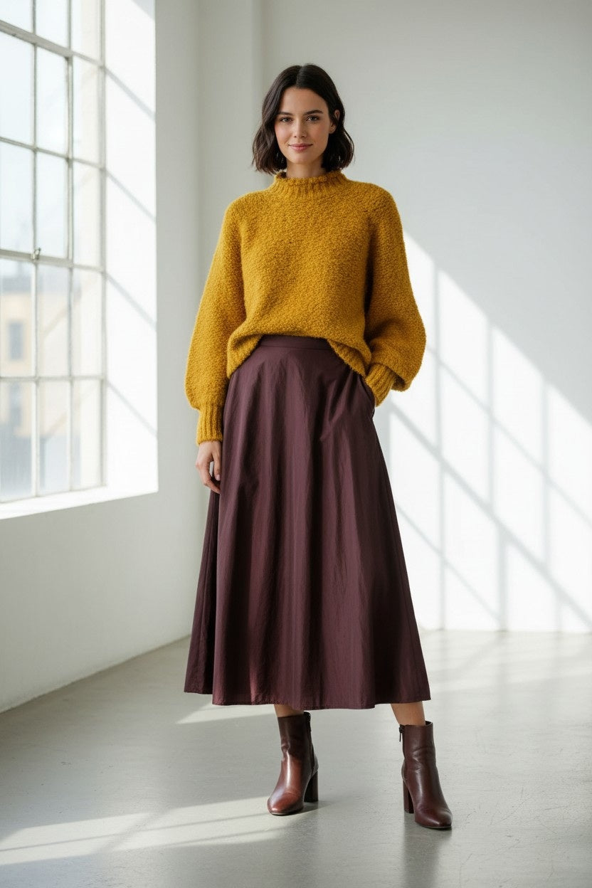 A woman stands in a sunlit room wearing a yellow sweater, the VICARIO CINQUE GRANADA Taffeta Midi Skirt in Bordeaux, and brown ankle boots. Sunlight pours through large windows, casting shadows across the light floor and white walls.