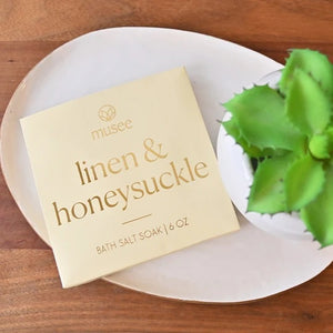 A package labeled “MUSEE - LINEN & HONESUCKLE MINI BATH SALT SOAK” from MUSEE rests on a white plate beside a green succulent on a wooden surface, highlighting exquisite natural bath products crafted with Pacific sea salt for the ultimate in relaxation.