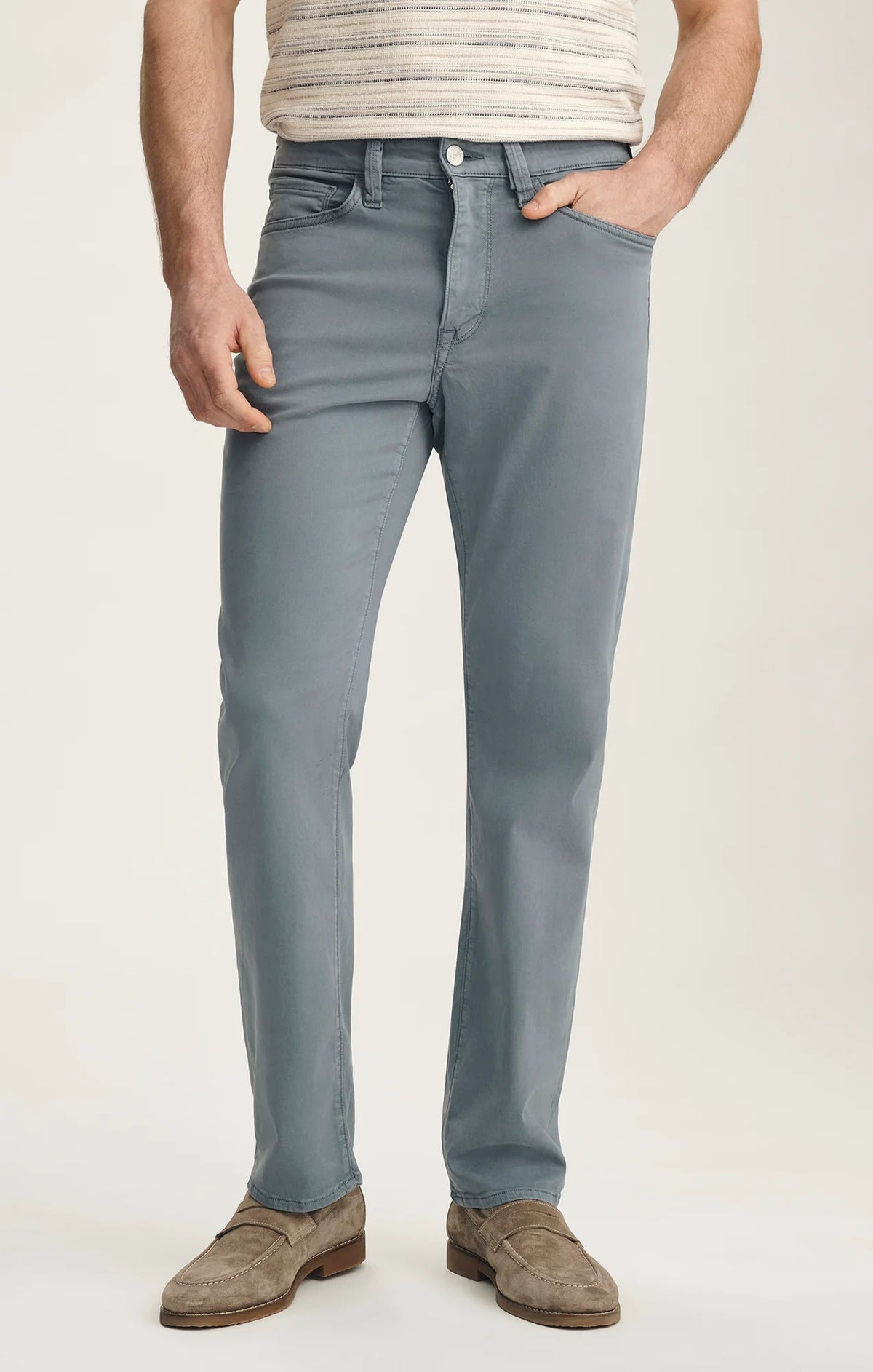 A man stands with his back to the camera against a plain light background, wearing a beige striped short-sleeve shirt, 34 HERITAGE Charisma Relaxed Straight Leg Pant in Flint Stone, brown suede shoes, and sunglasses.