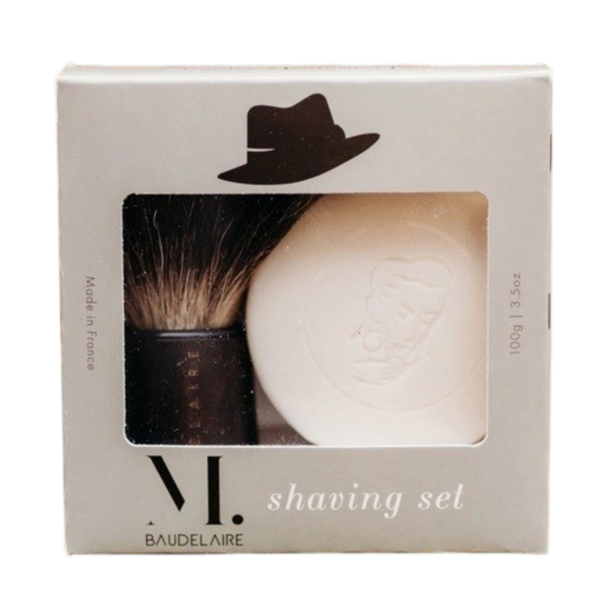 A hand holds the BAUDELAIRE - M.B. SHAVING SET bowl filled with foamy shaving soap, using the included badger bristle brush from BAUDELAIRE to whip up a rich lather.