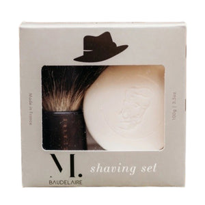 A hand holds the BAUDELAIRE - M.B. SHAVING SET bowl filled with foamy shaving soap, using the included badger bristle brush from BAUDELAIRE to whip up a rich lather.