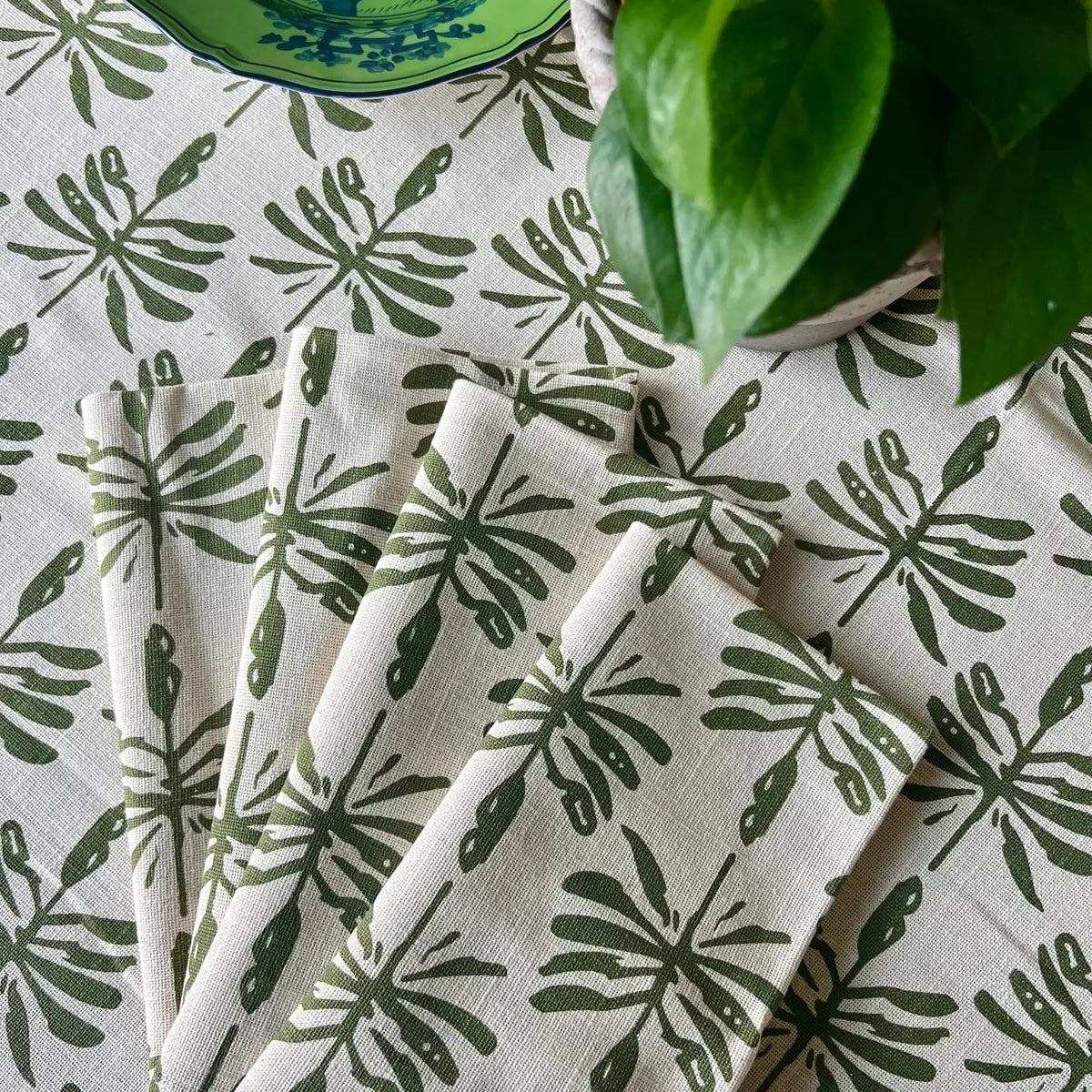 The CHRISTINA DICKSON HOME tablecloth features a green leaf pattern in cotton slub using handmade block print techniques, accompanied by folded napkins in the same design. A leafy plant and a decorative blue-patterned bowl on the tablecloth enhance its appeal.