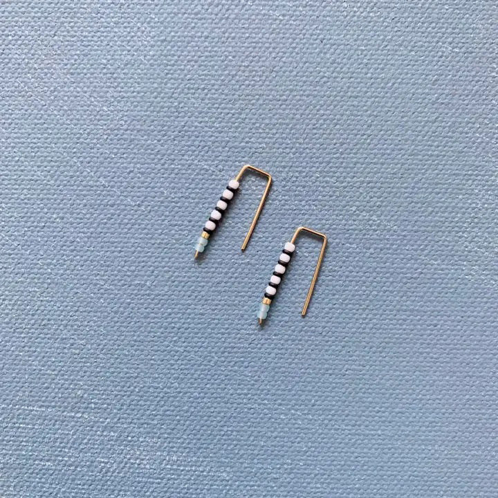 The ALICE RISE - LITTLE QUILL EARRINGS IN TURQUOISE by ALICE RISE, featuring minimalist 14k gold filled design and turquoise beads, are displayed on a textured light blue background—perfect for handmade jewelry lovers.