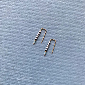 The ALICE RISE - LITTLE QUILL EARRINGS IN TURQUOISE by ALICE RISE, featuring minimalist 14k gold filled design and turquoise beads, are displayed on a textured light blue background—perfect for handmade jewelry lovers.