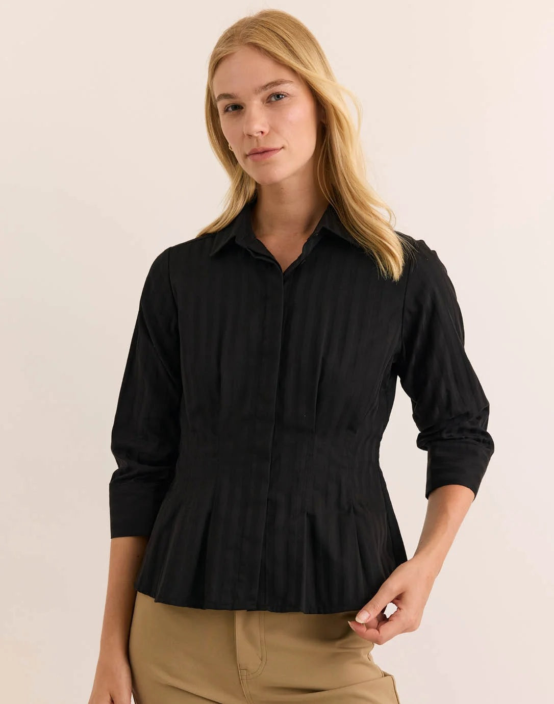 A woman is pictured wearing the HINSON WU - MARTINA 3/4 SLEEVE SHIRT IN CLASSIC STRIPE BLACK, a black button-up blouse with vertical stripes from HINSON WU, paired with tan pants. The shirt is crafted from breathable 100% cotton for all-day comfort. She stands with arms gently crossed and holds her collar, her face partially visible.