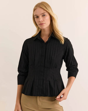 A woman is pictured wearing the HINSON WU - MARTINA 3/4 SLEEVE SHIRT IN CLASSIC STRIPE BLACK, a black button-up blouse with vertical stripes from HINSON WU, paired with tan pants. The shirt is crafted from breathable 100% cotton for all-day comfort. She stands with arms gently crossed and holds her collar, her face partially visible.