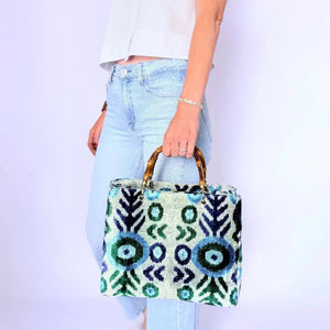 A person in light blue jeans and a white sleeveless top holds the BALDIZ AVERY Bag in Aqua Vine with bamboo handle. This stylish BALDIZ handbag’s blue, green, and white geometric design makes it fashionable and functional for any outing.