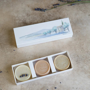 A hand holds a L'AUGUSTE PROVENCE handcrafted French soap bar and rubs it on a person's leg, creating a creamy lather that suggests an exfoliating skincare routine using artisanal soaps from L'AUGUSTE PROVENCE.