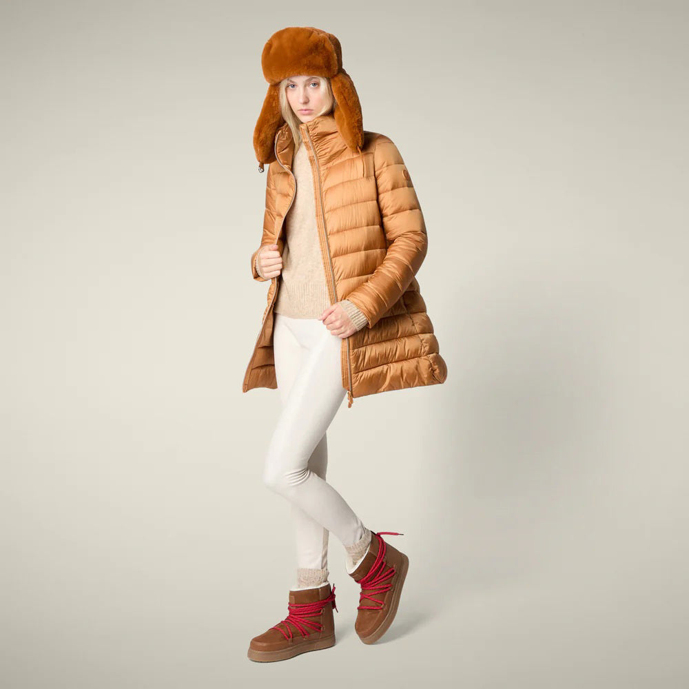 A woman in the SAVE THE DUCK - LYDIA PUFFER COAT by SAVE THE DUCK, paired with a white dress, maroon knee-high boots, and holding a fluffy light pink clutch, stands against a plain beige background.