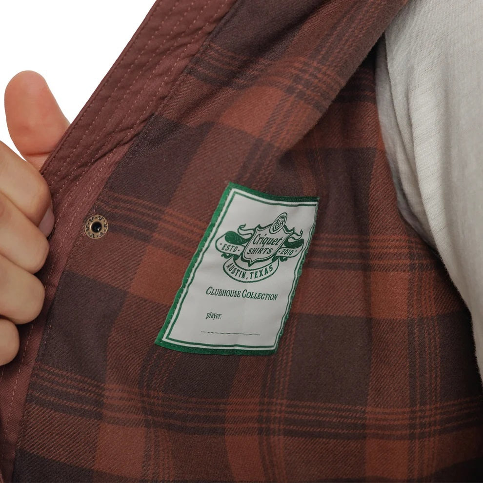 Close-up of the interior of a brown plaid CRIQUET - WAXED RANCH JACKET IN BOURBON, showing a white “Clubhouse Collection” brand label with a blank line for a player’s name.