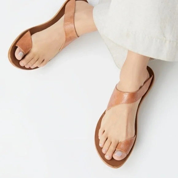 A person wearing light beige pants and tan leather sandals with a unique twisting strap design, photographed from above against a white background.