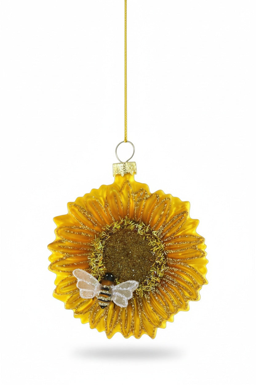 The CODY FOSTER SUNFLOWER WITH BEE ORNAMENT features gold glitter accents and a charming bee, making it an ideal collector’s piece or gift for design lovers, displayed hanging by a gold string against a white background.
