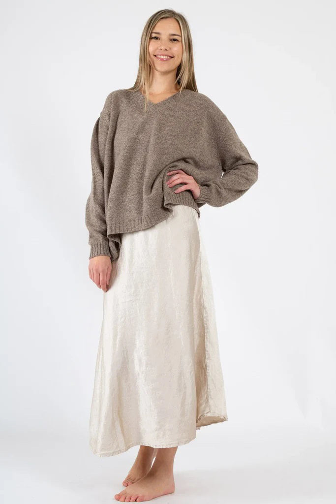 A person with long hair is smiling while wearing an oversized brown sweater and the CP SHADES - TANYA SATIN SLUB SKIRT, a flowing ankle-length cream skirt. They are standing barefoot against a plain white background.