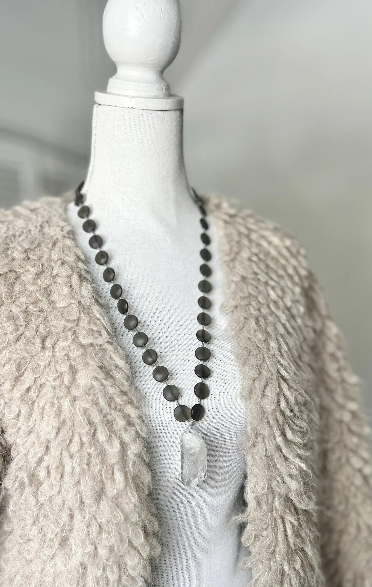 The ANN LIGHTFOOT Quartz and Smokey Glass Disc Pendant Necklace showcases round, dark smokey glass beads paired with a striking clear quartz pendant. Elegantly displayed on a white background, this 30-inch necklace by ANN LIGHTFOOT blends refined sophistication with natural allure.