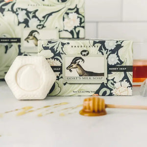 A BAUDELAIRE GOATS MILK HONEY SOAP 3.5OZ 2 BAR GIFT BOX features a floral, goat-illustrated box labeled "Honey Soap." Inside are two hexagonal, white moisturizing bars with embossed designs—ideal for sensitive skin.