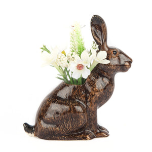 The QUAIL DESIGNS QUAIL CERAMICS - HARE BUD VASE is a hand-painted stoneware vase shaped like a rabbit, featuring a brown and black finish. It displays small white flowers and greenery in the opening on its back against a plain white background.