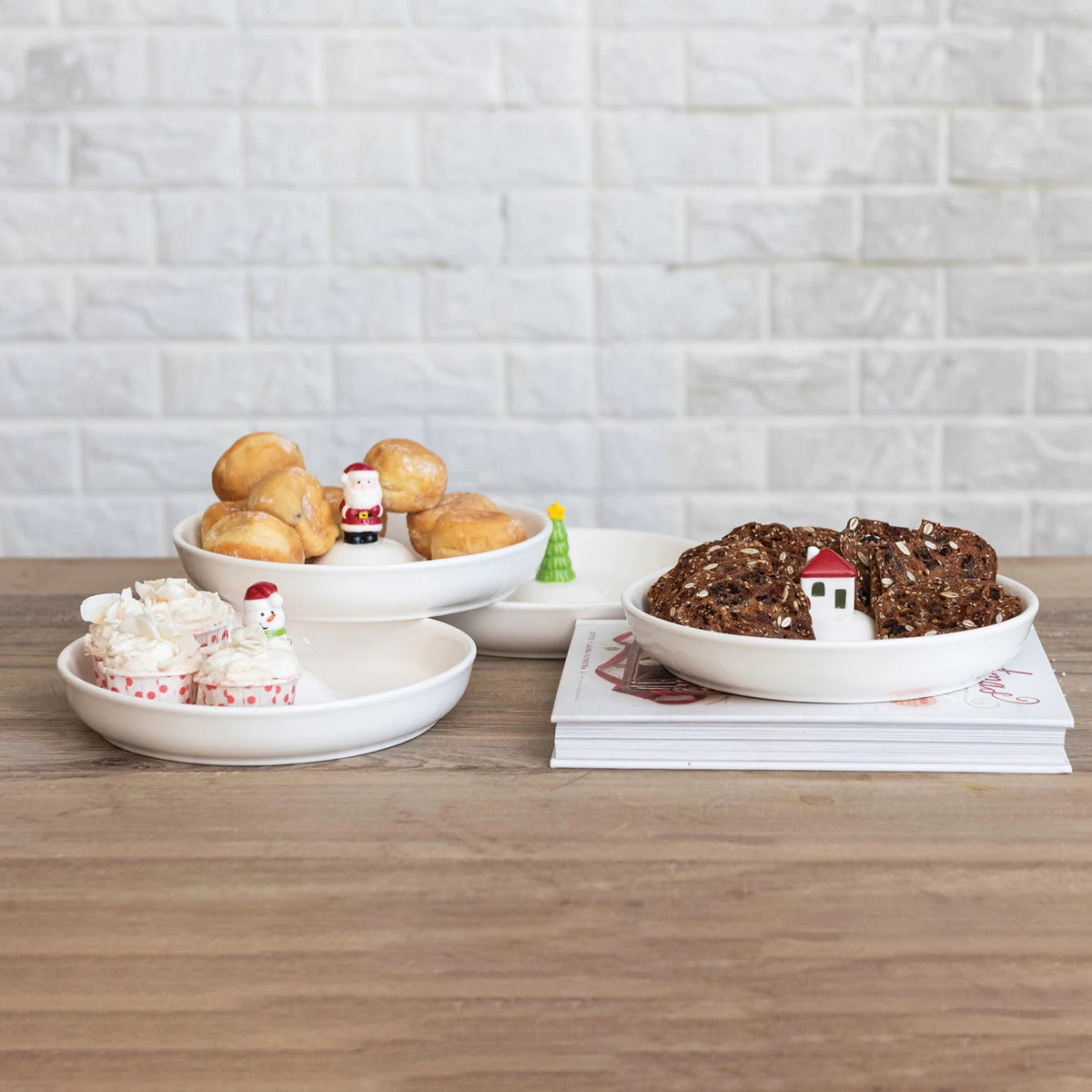 Three CREATIVE COOP STONEWARE DISHES WITH HOLIDAY FIGURES hold cream puffs, mini cupcakes, and crispbread on a wooden table, two stacked with Santa and Christmas tree décor, set against a brick wall backdrop.