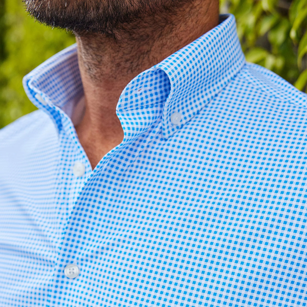 A man with short dark hair and a trimmed beard, wearing the COLLARS AND CO - Oxford Button Down Collar Apex Performance Dress Shirt in Micro Blue Gingham, holds open a glass door and looks left in a bright, modern space.