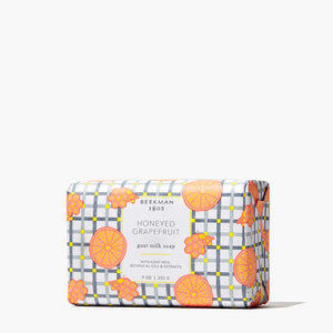 The BEEKMAN - STA ELEMENTS HONEYED GRAPEFRUIT 3.5 SOAP is a rectangular exfoliating goat milk bar wrapped in white paper with a gray checkered pattern, featuring orange grapefruit slices and honeycomb illustrations for a natural glow.