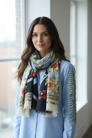 The STORIATIPIC ARIANE wool scarf with embroidery is adorned with a floral and vine pattern in red, green, blue, cream, and black tones. It features light blue edges and a soft, slightly fringed border. The embroidered scarf is loosely knotted against a white background.