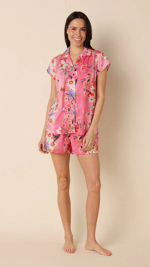 A woman smiles barefoot against a beige background, wearing CAT'S PAJAMAS - SPRING FLING VOILE SHORT SET: a lightweight cotton voile pajama with a colorful floral print and dark piping, perfect for comfortable women's sleepwear.