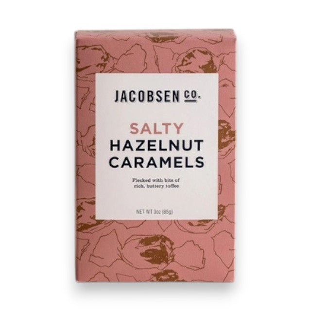 A pink JACOBSEN SALT - SALTY HAZELNUT CARAMELS BOX by JACOBSEN SALT CO. sits on a light textured surface, featuring a modern design with subtle hazelnut patterns and hints of buttery toffee made from premium hazelnuts.