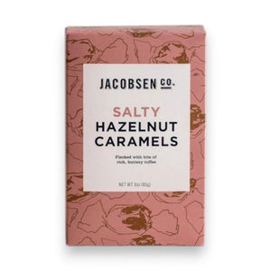 A pink JACOBSEN SALT - SALTY HAZELNUT CARAMELS BOX by JACOBSEN SALT CO. sits on a light textured surface, featuring a modern design with subtle hazelnut patterns and hints of buttery toffee made from premium hazelnuts.