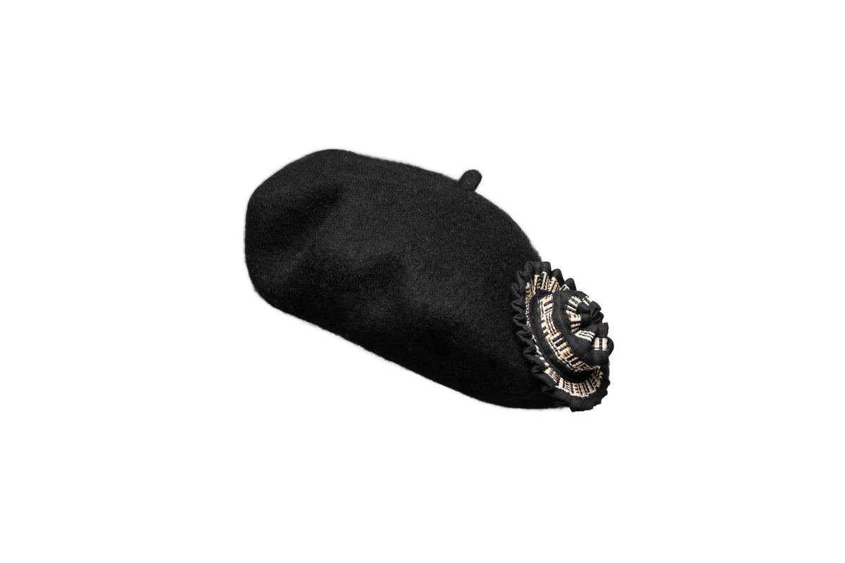 The LORNA MURRAY - THE AVENUE BERET from LORNA MURRAY is a black beret crafted from premium Australian wool. It features a detailed, black and silver emblem on the side, which resembles an intricately decorated crest or badge, adding an elegant touch to its classic style. The plain white background emphasizes the sophistication of this stunning accessory.