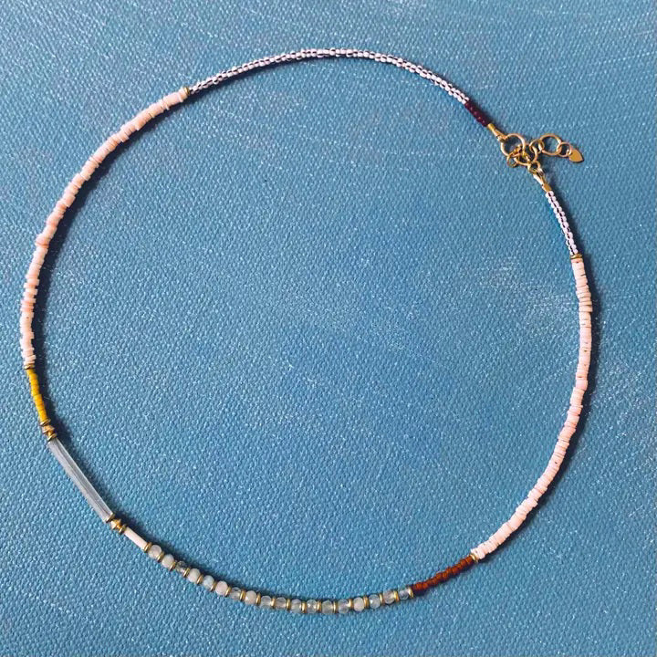 The ALICE RISE - AQUAMARINE NECKLACE by ALICE RISE features small multicolored beads and a gold clasp, handmade in North Carolina, and is displayed in a circle on a textured blue background.