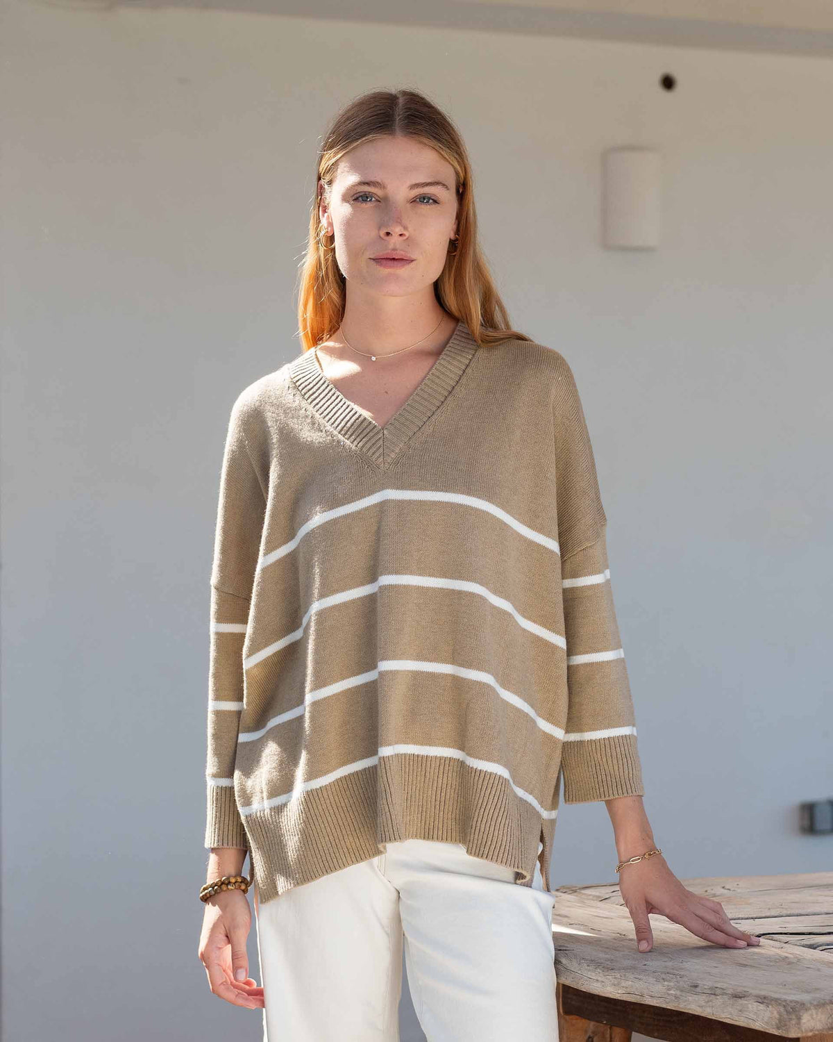 A woman wears the MERSEA - MONTAUK V-NECK SWEATER by MERSEA, a white oversized soft knit with blue and green striped collar, cuffs, and hem, ribbed v-neckline, styled over denim shorts and a light pink shirt.