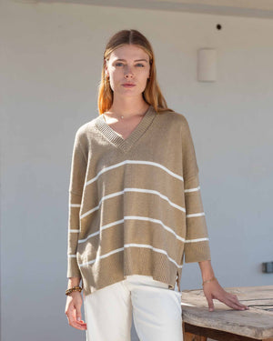 A woman wears the MERSEA - MONTAUK V-NECK SWEATER by MERSEA, a white oversized soft knit with blue and green striped collar, cuffs, and hem, ribbed v-neckline, styled over denim shorts and a light pink shirt.