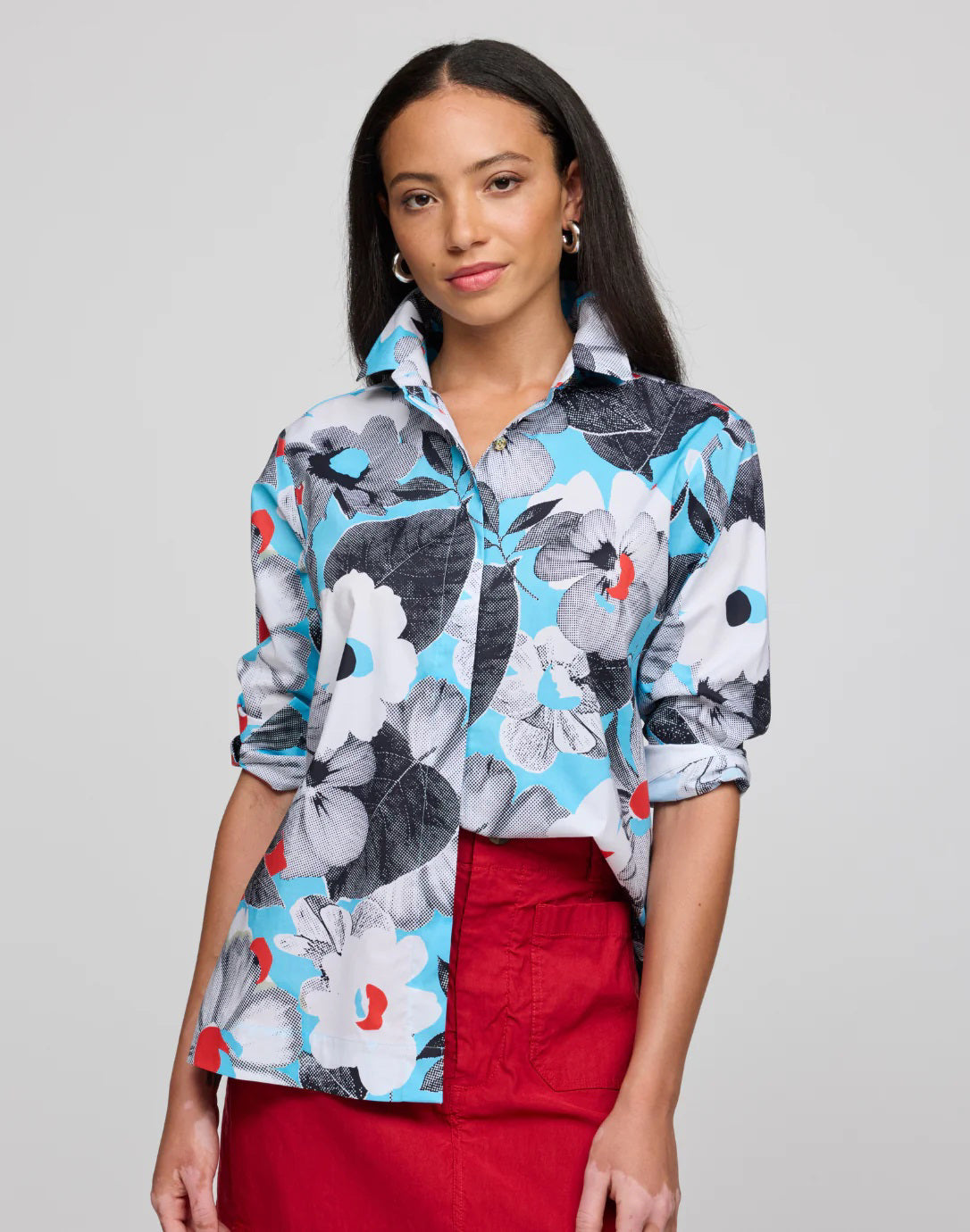 A woman is wearing the HINSON WU - HALSEY SHIRT IN POP ART FLOWER, featuring a colorful floral pattern in blue, white, and black. She rolls up its sleeves and pairs it with a red skirt, complemented by her long straight hair and hoop earrings against a plain background.