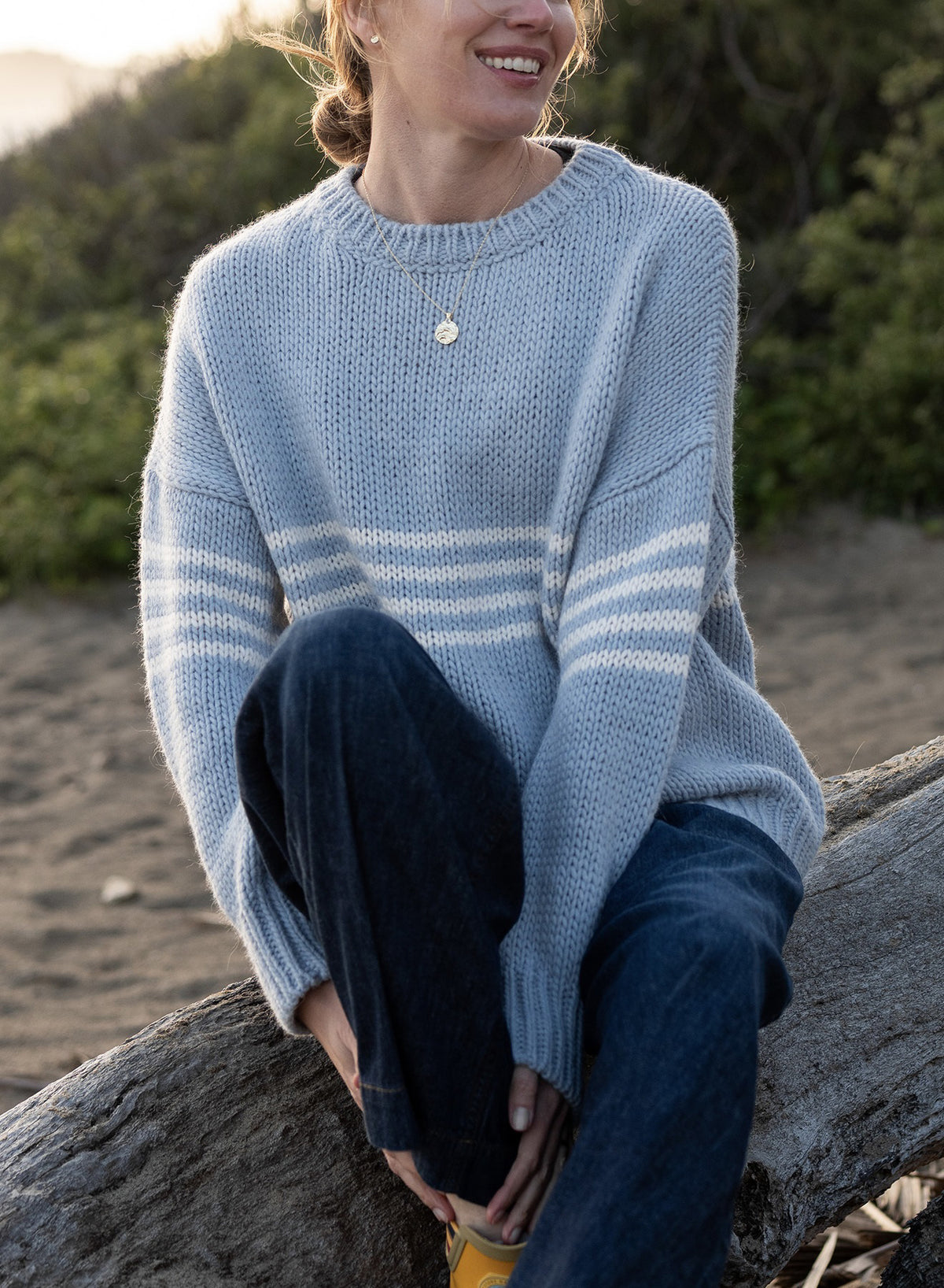 Smiling outdoors, a person wears the MERSEA - SEACOAST STRIPE SWEATER—a light blue knit with white sleeve stripes—paired with dark jeans and a gold necklace. Green foliage and sandy ground enhance the cozy, coastal vibe.