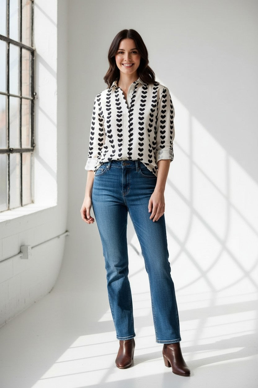 A person wearing the EMILY PHILLIPS - BLACK HEARTS GIRLFRIEND BUTTON DOWN SHIRT, a sustainably made white cotton top featuring rolled sleeves and vertical rows of large black heart patterns.