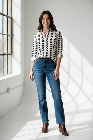 A person wearing the EMILY PHILLIPS - BLACK HEARTS GIRLFRIEND BUTTON DOWN SHIRT, a sustainably made white cotton top featuring rolled sleeves and vertical rows of large black heart patterns.