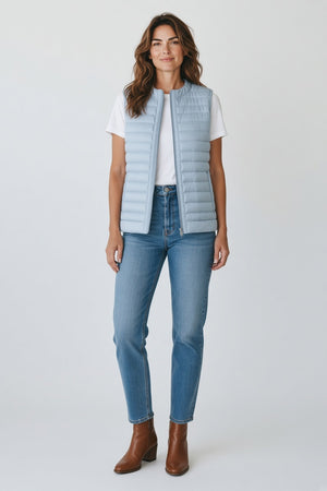 An older woman with short gray hair wears the SAVE THE DUCK - MARA PUFFER VEST over a white t-shirt and beige pants, standing against a light background—perfect for spring–summer layering from SAVE THE DUCK.