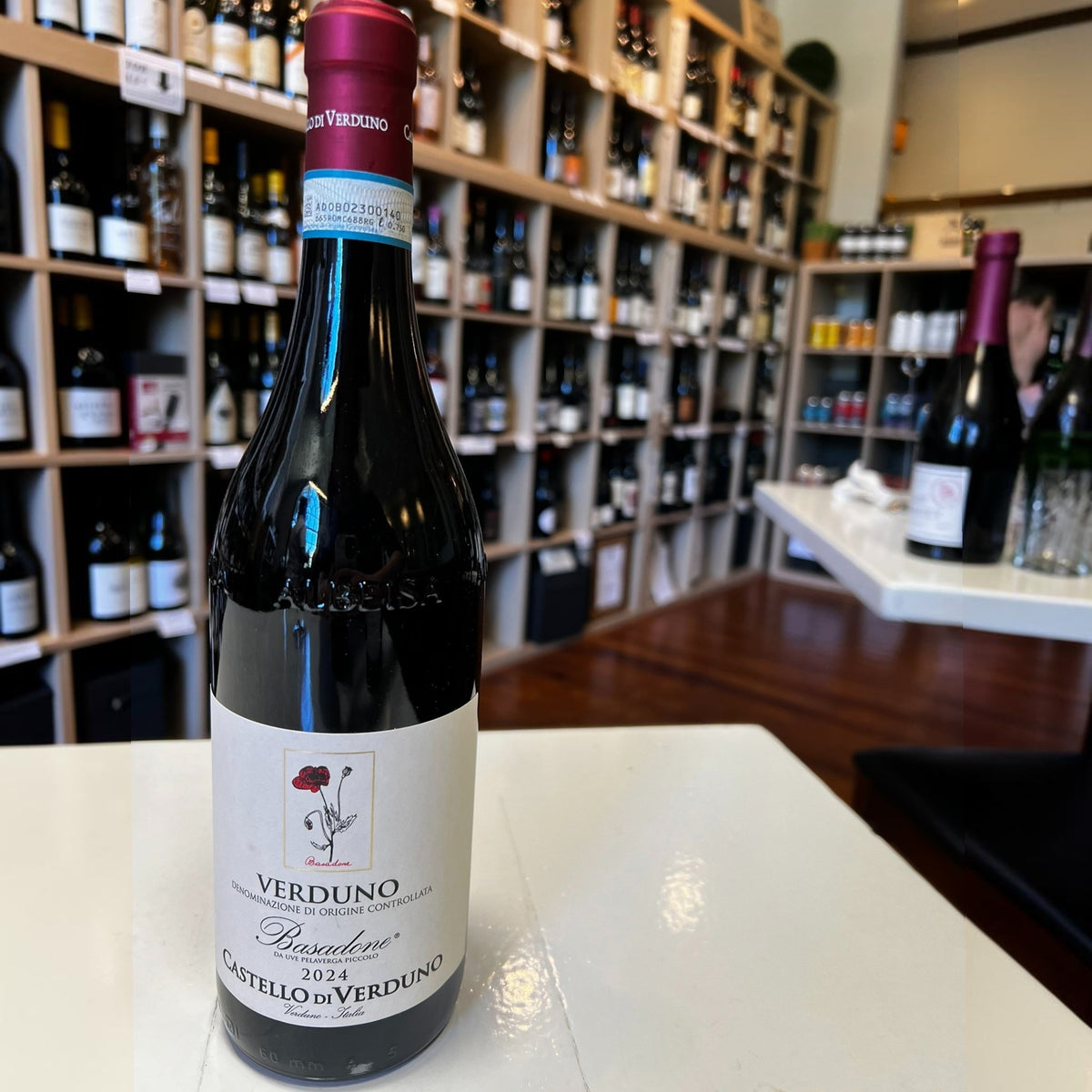 A bottle of CASTELLO DI VERDUNO ‘BASADONE’ PELAVERGA 2024 red wine with a label featuring an illustration of a red flower, sitting on a white table in a wine shop.