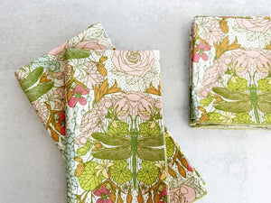 A neat stack of DRAGONFLY GARDEN COCKTAIL NAPKINS by DOT AND ARMY, made from 100% cotton. These super soft napkins feature a vibrant floral print with pink and white flowers, green leaves, and a large green dragonfly in the center, all set on a light gray surface.