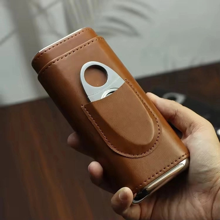 The AMERICAN LEATHER GOODS Leather Cigar Case with wood-lined humidor and cutter in brown sits on a wooden surface, while a black case rests behind. Leaf shadows appear on the wall in the background.