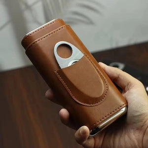 The AMERICAN LEATHER GOODS Leather Cigar Case with wood-lined humidor and cutter in brown sits on a wooden surface, while a black case rests behind. Leaf shadows appear on the wall in the background.