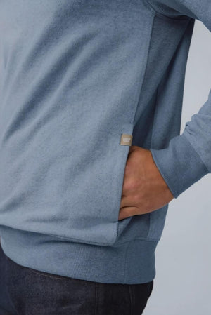 A man with short light brown hair stands in profile, wearing the MACK WELDON Ace 1/2 Zip Sweater in Herringbone and dark pants, one hand in his pocket, against a plain light gray background.