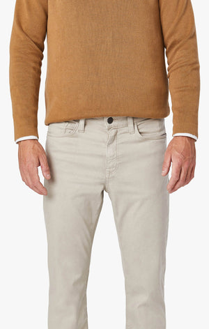A bearded man stands against a plain background, wearing a tan blazer, a brown sweater over a white shirt, and 34 HERITAGE - CHARISMA RELAXED STRAIGHT LEG PANT IN DAWN TWILL. He adjusts his blazer sleeve with one hand.