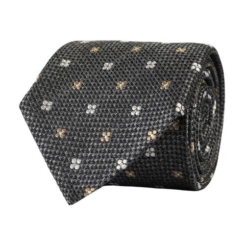 The AMANDA CHRISTENSEN - FLORAL JACQUARD WOVEN SILK TIE IN GREY MELANGE, elegantly rolled and displayed on a white background, features a sophisticated dark gray mélange base adorned with small white and beige floral patterns—an exquisite addition to any men’s accessories collection by AMANDA CHRISTENSEN.