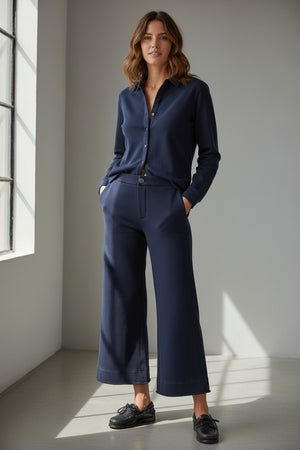 A person stands outdoors facing away, wearing the FRANK & EILEEN Riley Trouser Sweatpant in Triple Fleece Classic Navy paired with black shoes, one hand resting by their side near a wooden wall and a bench—a relaxed look inspired by FRANK & EILEEN’s California Heritage Collection.