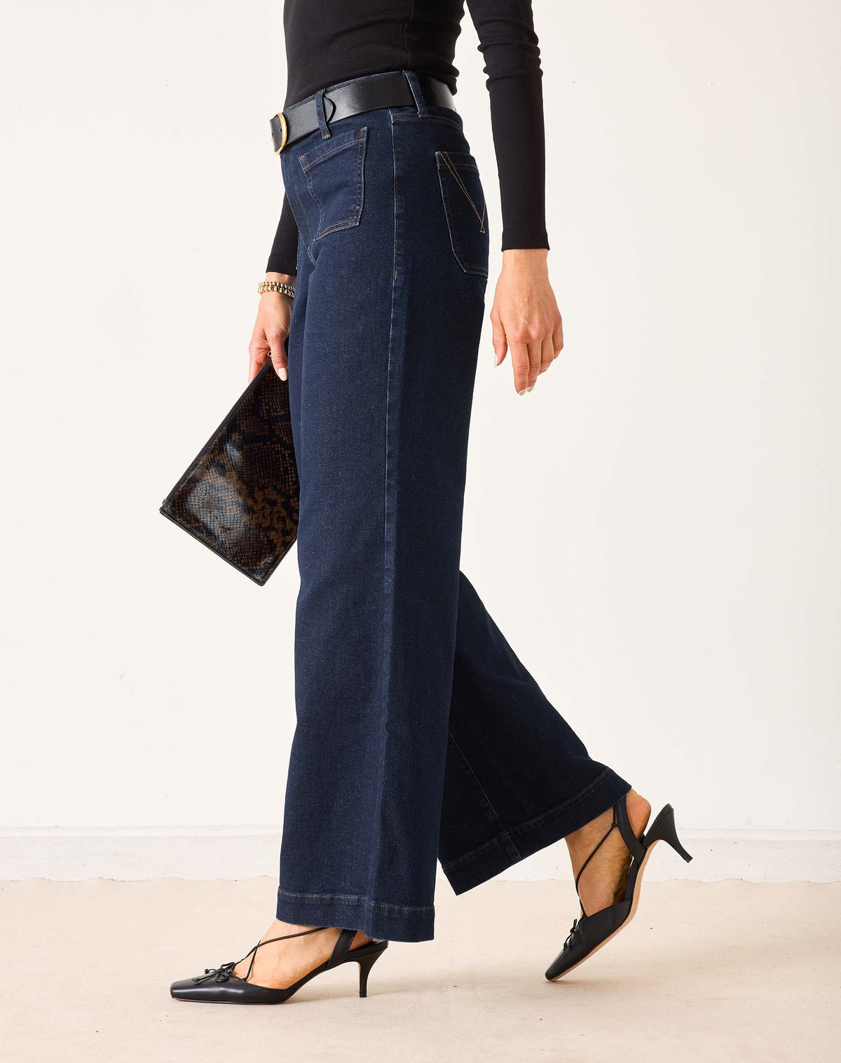 A person wearing MERSEA - SAILOR WIDE LEG JEANS IN RINSE by MERSEA, styled with a black top, black belt, and black heeled sandals, stands with their back to the camera against a plain light background.