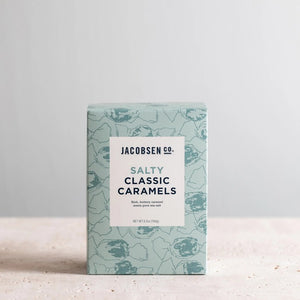 A light blue box of JACOBSEN SALT CO. JACOBSEN SALT - SALTY CARAMELS BOX, featuring a patterned design and product label on the front, rests on a light surface.