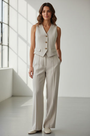 A person with long dark hair models the SPANX - CAREFREE CREPE VEST, a sleeveless, light beige tailored vest with gold buttons, paired with matching high-waisted trousers, standing hands in pockets against a plain white background.